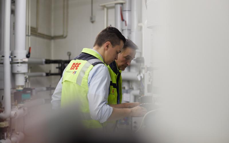 zwei Ingenieure vor Heizungsanlage mit gelben Schutzwesten mit BFE Logo